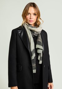 Zwarte jas met leren revers, gelaagde grijze en groene sjaal met een gestreept patroon. Zachte texturen en vloeiende stof.