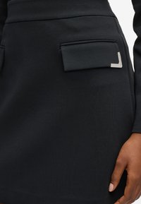 Gonna nera con tasche a pozzetto e dettagli angolari argentati, indossata con un top coordinato e mano visibile con smalto neutro.