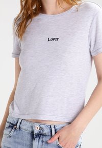 Camiseta gris de manga corta con cuello redondo, que presenta la palabra "Lover" en bordado negro en el pecho. Tejido suave y corte relajado.