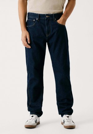 Dark blue denim jeans with a straight-leg fit, featuring contrasting orange stitching, side pockets, and a button closure. Paired with white sneakers.