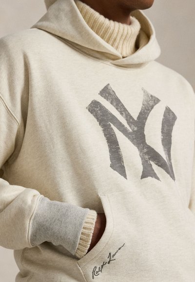 Ανοιχτό μπεζ φούτερ με κουκούλα, που διαθέτει γραφικό NY σε σκούρο γκρι μπροστά. Περιέχει τσέπη καγκουρό, ριμπ μανσέτες και ένα διακριτικό λογότυπο κοντά στην τσέπη.