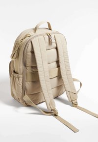 Beige quiltad ryggsäck med vadderade remmar, handtag upptill, dragkedjeslås och sidoficka. Mjukt material och minimalistisk design.