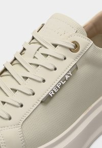 Gros plan sur une basket beige en tissu texturé, avec des lacets assortis, des œillets métalliques et le logo "REPLAY" sur le panneau latéral.