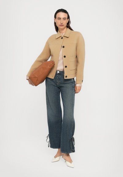 Frau in einer camel farbenen, geknöpften Jacke, blauen, weiten Jeans mit Bindungen, weißen Slingback-Absätzen und die eine große braune Lederhandtasche trägt.