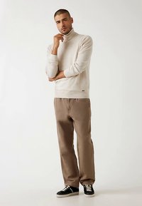 Béžový rolák v kombinácii so svetlohnedými nohavicami a čiernymi teniskami, ktorý vyniká priliehavým strihom a hladkou textúrou po celej ploche.