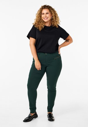 Musta lyhythihainen T-paita yhdistettynä vihreisiin kapeisiin housuihin, joissa on vetoketjullinen tasku, sileä pintakuvio ja klassinen siluetti. Mustat loafersit jaloissa.
