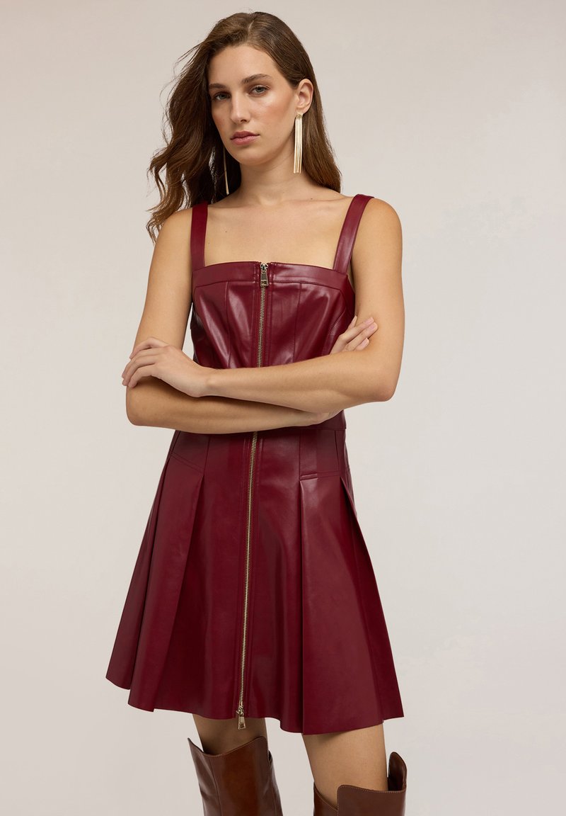 Donna con lunghi capelli mossi che indossa un vestito in pelle bordeaux senza maniche con cerniera frontale e stivali marroni al ginocchio, con le braccia incrociate.