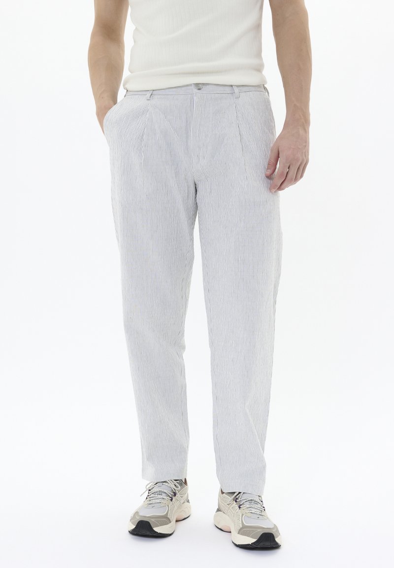 Pantalons légers à rayures bleues et blanches, offrant une coupe ajustée, des plis à l'avant et des poches latérales. Accompagnés de baskets neutres.