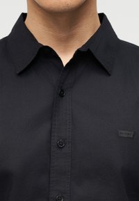 Άνδρας φοράει μαύρο πουκάμισο με κουμπιά και γιακά, με μικρό λογότυπο Levi's στο στήθος.