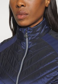 Námornická prošívaná bunda s vysokým límcem, s páskovým vzorem, hladkým povrchem a stříbrným zipem se zaobleným táhlem.