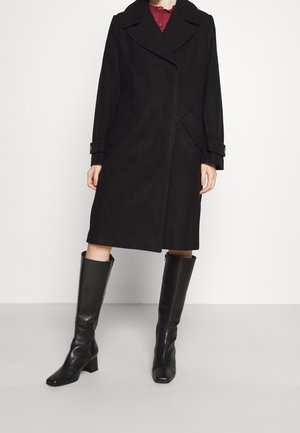 Femme portant un manteau noir mi-long aux genoux avec un large col et des bottes noires montantes, debout devant un fond clair uni.