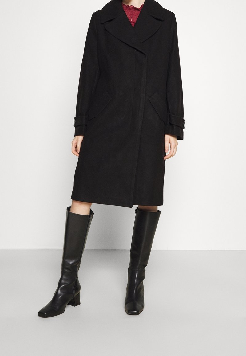 Femme portant un manteau noir mi-long aux genoux avec un large col et des bottes noires montantes, debout devant un fond clair uni.