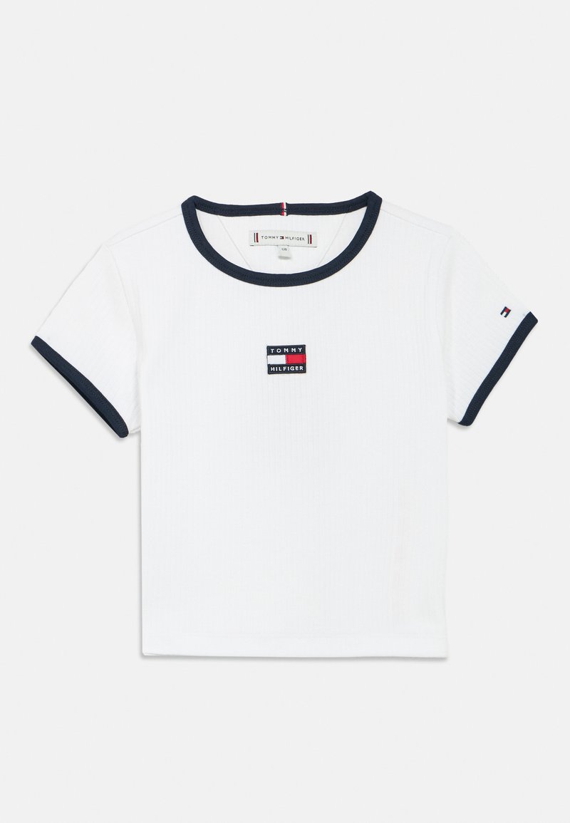 Tricou alb cu dungi, cu tiv bleumarin la guler și mâneci. Prezintă o mică etichetă logo dreptunghiulară, în roșu, alb și albastru, pe piept.