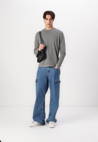 Šedá dlouhá košile, oversized modré džíny s cargo kapsami, bílé tenisky a černá crossbody taška s minimálním kovovým příslušenstvím.