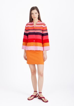 Frau trägt eine bunte gestreifte Strickjacke in Weiß-, Rot-, Orange- und Lilatönen, kombiniert mit einem orangen Rock und roten Sandalen.