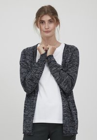 Cardigan tricoté en bleu marine foncé et gris avec un motif texturé. Ouvert sur le devant, manches longues, porté par-dessus un t-shirt blanc uni.