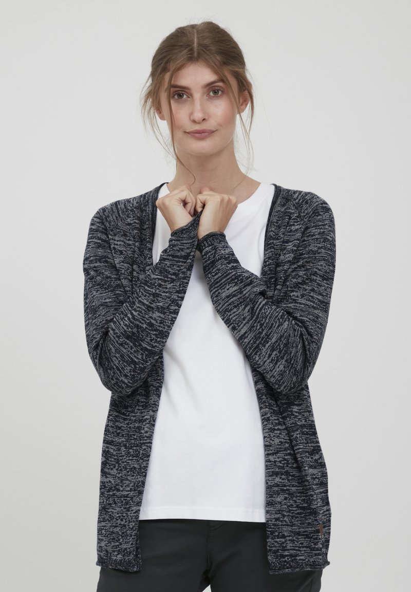 Cardigan tricoté en bleu marine foncé et gris avec un motif texturé. Ouvert sur le devant, manches longues, porté par-dessus un t-shirt blanc uni.