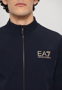 Temno modra zip-up jakna z visokim ovratnikom in zlatim logotipom na levem prsnem delu. Gladko blago in standardna zasnova.