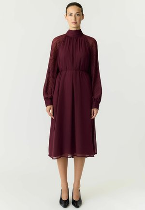 Femme debout dans une robe midi bordeaux à manches longues transparentes texturées et chaussures noires à talons hauts sur un fond uni.