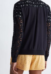 Černá bunda s krajkovými detaily na rukávech, s hladkým zadním dílem a manžetami. Spárovaná s žlutými šortkami s květinovým krajkovým vzorem.