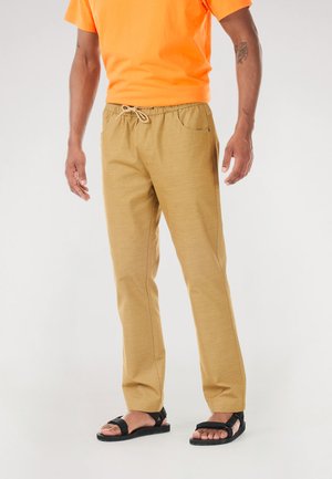 Beige Hose aus einem weichen, strukturierten Stoff mit elastischem Bund und Kordelzug, kombiniert mit einem leuchtend orangefarbenen T-Shirt und schwarzen Sandalen.