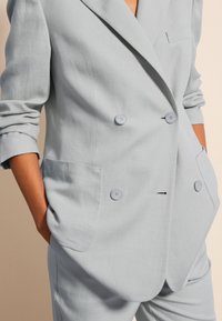 Blazer de lino azul claro de doble botonadura con solapas en pico, dos botones y un único bolsillo en el pecho. Presenta mangas remangadas y bolsillos laterales.