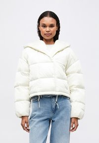 Γυναίκα που φοράει ένα λευκό μπουφάν με κορδόνια στο τελείωμα και ανοιχτό μπλε τζιν ψηλόμεσα, σε ουδέτερο φόντο.