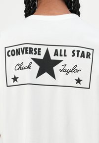 Balta kokvilnas T-krekls ar melnu grafiku uz muguras, kurā ietilpst teksts "CONVERSE ALL STAR" un centrālais melns zvaigznes dizains.