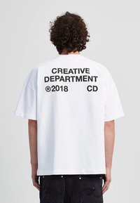Weißes Baumwoll-T-Shirt mit lockerer Passform, auf dem Rücken ist großer schwarzer Text aufgedruckt: "CREATIVE DEPARTMENT ® 2018 CD." Kein Vorderdesign.