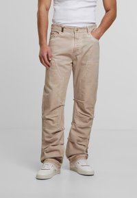 Mies, joka on pukeutunut vaaleisiin cargo-housuihin, valkoisiin tennareihin ja valkoiseen hihattomaan paitaan, seisoo yksi käsi taskussa ja toinen käsi rentona.