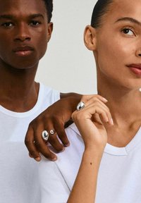 Due persone in magliette bianche mostrano anelli metallici. Un anello è ovale con una finitura lucida, mentre l'altro è più piccolo e rotondo.