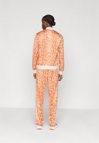 Persona ar pītām matiem stāv, ģērbusies oranžas un bēšas dzīvnieku raksta jaku un biksēs, ar baltām sporta kurpēm, skatoties prom uz vienkārša fona.