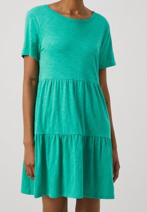 Kobieta ubrana w zieloną, krótko rękawową sukienkę warstwową z okrągłym dekoltem, stojąca na tle jednolitego jasnego tła.