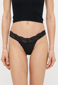 String en coton et dentelle noire avec une bordure en dentelle florale à la taille, un petit nœud décoratif à l'avant, et un design minimaliste.