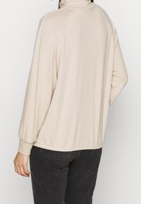 Haut beige à manches longues avec un col montant et des poignets élastiqués. Tissu doux avec une coupe décontractée. Détail notable des coutures latérales.