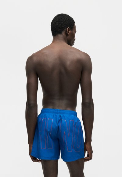 Άνδρας χωρίς μπλούζα με σκουρόχρωμο δέρμα, που φορά μπλε μαγιό σορτς με μεγάλα περιγραμμικά γράμματα, στέκεται με την πλάτη σε ένα απλό ανοιχτό φόντο.