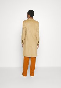 Cappotto beige con vestibilità su misura, caratterizzato da un colletto a punte e polsini con bottoni, abbinato a pantaloni arancioni a gamba larga e sneakers.