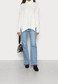 Suéter blanco de punto suelto con cuello alto, combinado con jeans de campana azul claro. Botines negros con tacón y un pequeño bolso negro acentúan el atuendo.