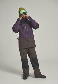 Snow jacket and pants in purple, brown, and black. The jacket features zippers, waterproof fabric, and a high collar. Green goggles and a knit hat.