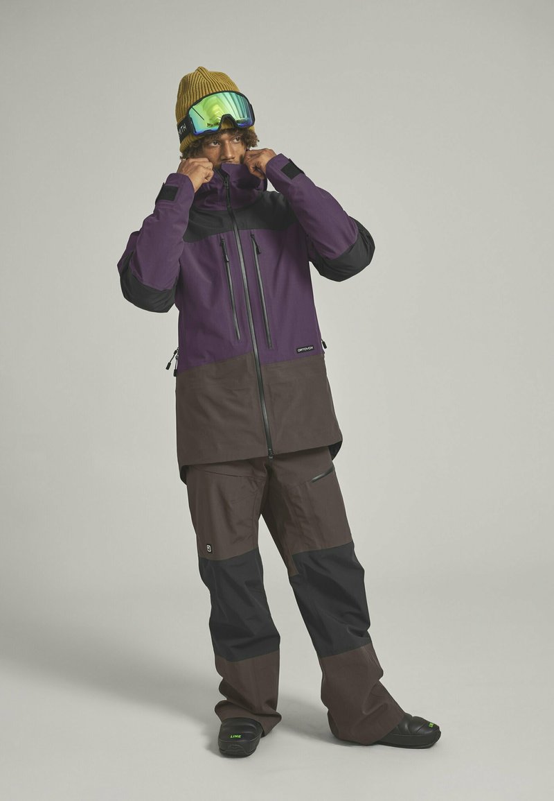 Snow jacket and pants in purple, brown, and black. The jacket features zippers, waterproof fabric, and a high collar. Green goggles and a knit hat.