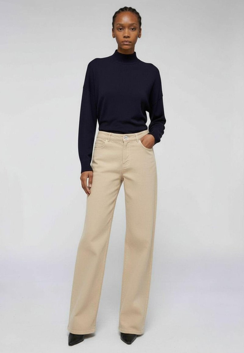 Pantalones de pierna ancha color beige, con costuras detalladas y un ligero brillo. Combinados con un suéter azul marino de cuello alto, con un ajuste relajado.