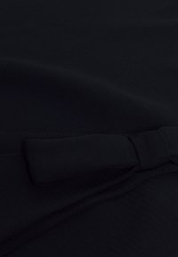 Černá látka s hladkou texturou, zdobená boční mašlí, prošitými okraji a jemným diagonálním vzorem.