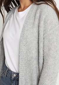 Cardigan grigio chiaro a maglia con apertura frontale, tessuto testurizzato, maniche ampie e vestibilità rilassata, indossato su una t-shirt bianca.