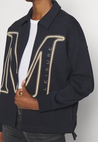 Giacca felpa blu navy con grande grafica illuminata della lettera "M" e la parola "LEISURE" sul davanti; vestibilità rilassata con tasche laterali.