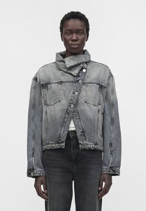 Osoba nosząca wyblakłą szarą kurtkę jeansową z asymetrycznym, zapinanym na guziki przednim panelem oraz wysokim kołnierzem, w połączeniu z ciemnoszarymi jeansami.