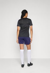 T-shirt de sport noir à manches courtes avec un dos structuré, associé à un short violet, des chaussettes blanches montantes et des chaussures de sport noires.
