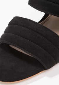 Černé semišové skládací sandály se dvěma širokými pásky, které mají měkkou texturu a otevřenou špičku. Přírodní kožená stélka s hnědými akcenty.