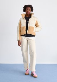 Beige en crème pufferjack met een pluche textuur, zwarte T-shirt, crème broek en roze sneakers met groene accenten.