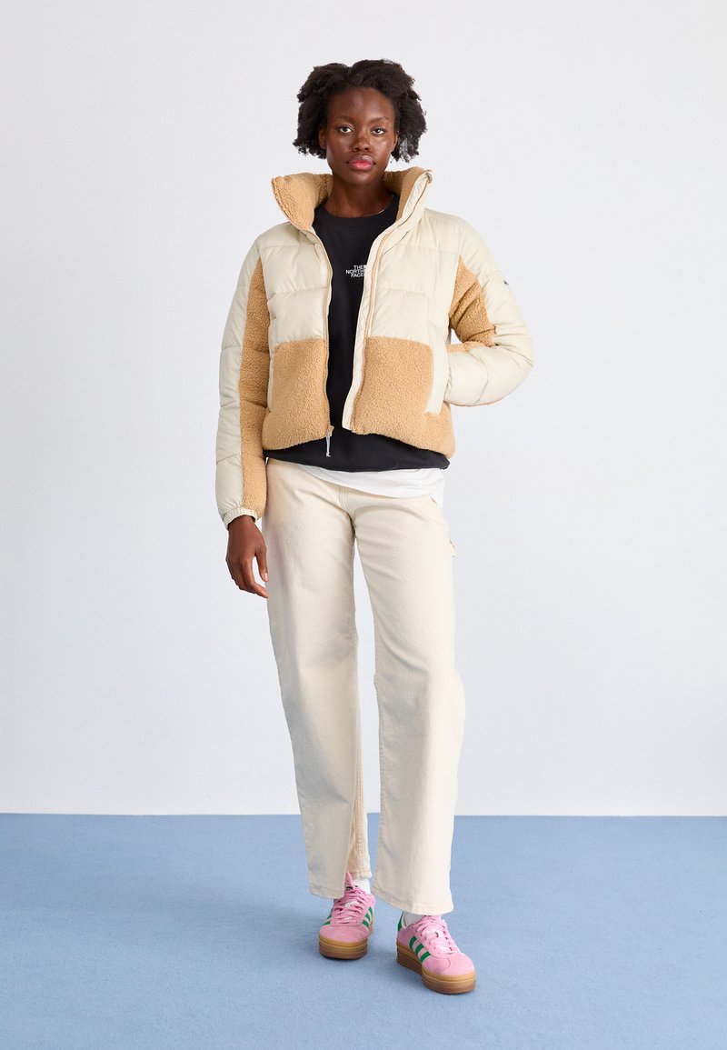 Beżowa i kremowa kurtka puchowa o puszystej fakturze, czarny t-shirt, kremowe spodnie oraz różowe sneakersy z zielonymi akcentami.