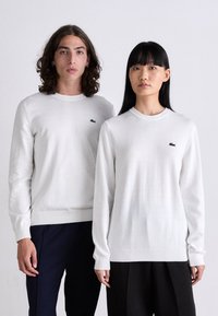 White long-sleeved sweaters featuring a round neckline and green embroidered logo on the chest, made from soft, textured fabric.